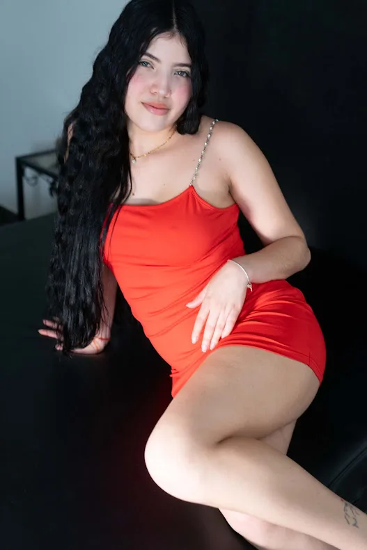 Roxy, Escort posiert sitzend im Kleid und ihrem langen schwarzen gewellten Haar in Velazquez24